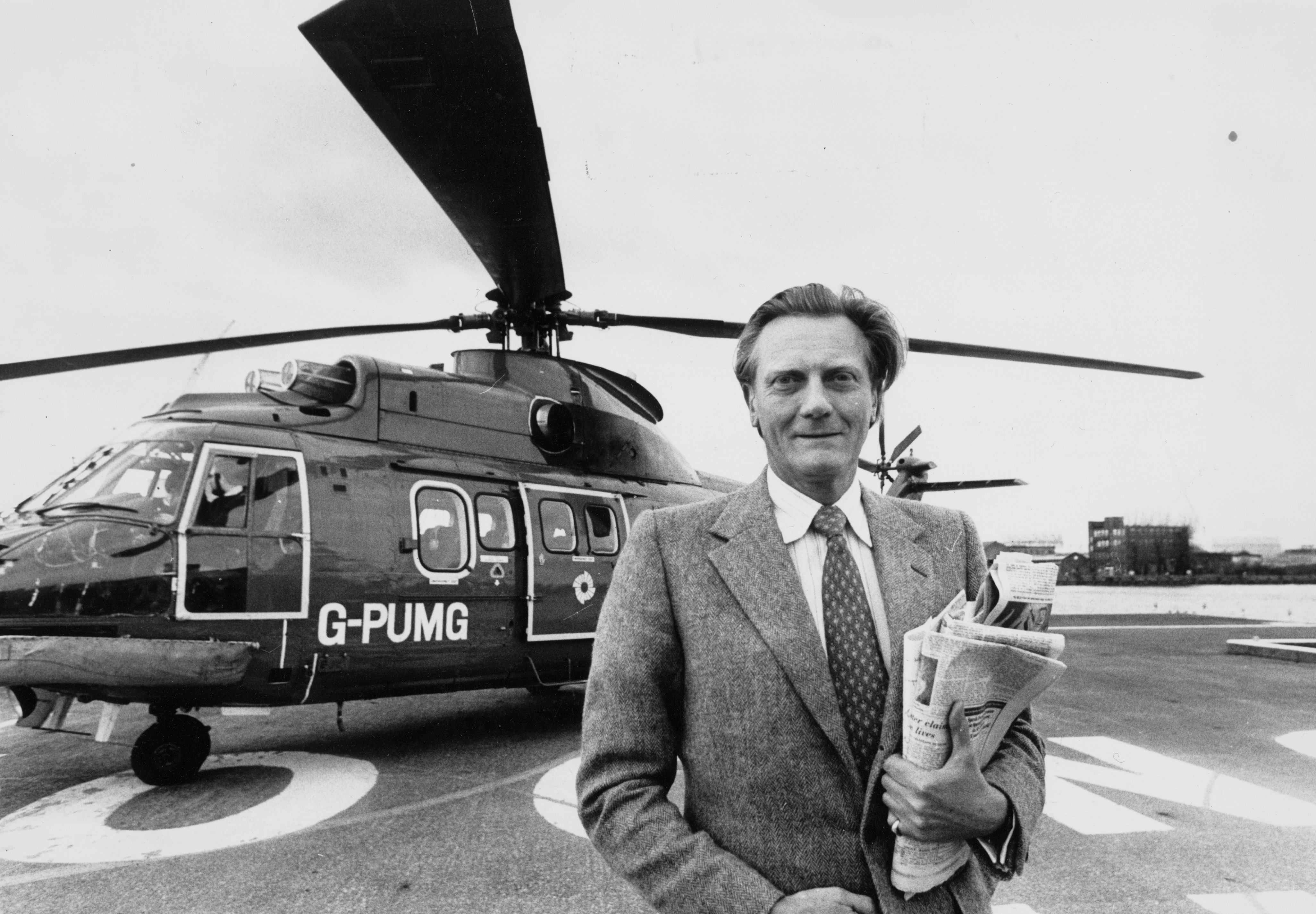 Michael Heseltine standing by a helicopter at a heliport, holding newspapers.