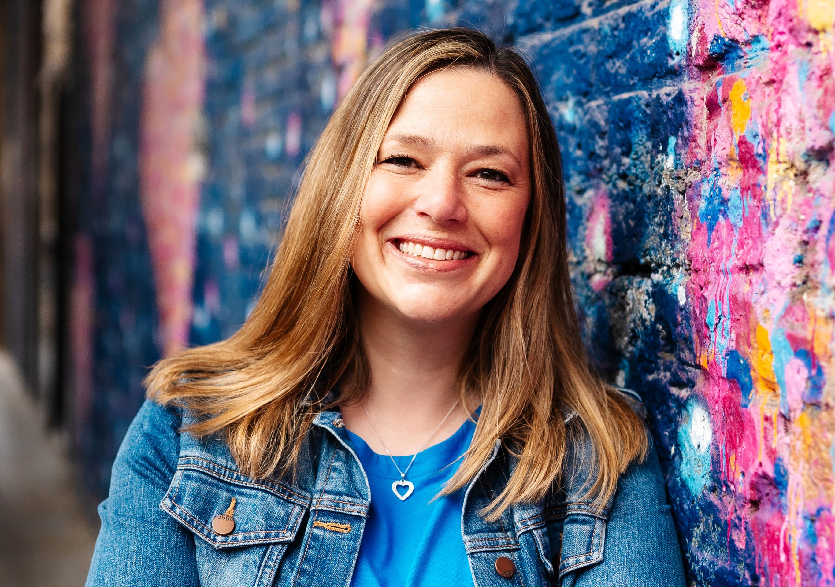 Woman in denim jacket smiling against colorful wall.